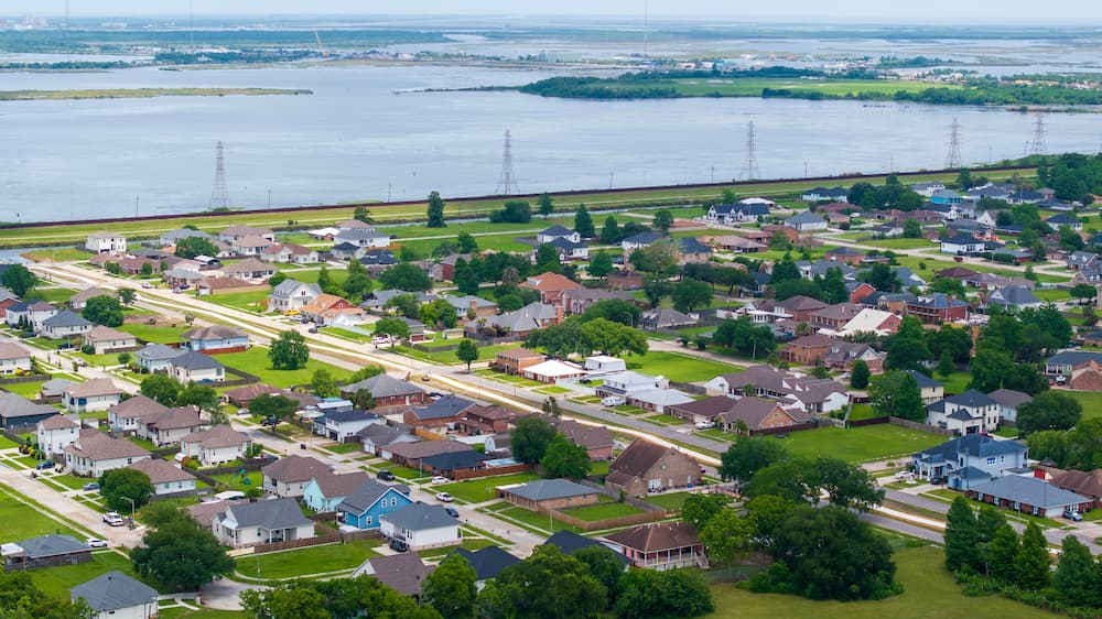 Aerial view of a dense suburban neighborhood in Louisiana near a large body of water, emphasizing the ongoing inspection and roof maintenance required due to high winds and humidity.