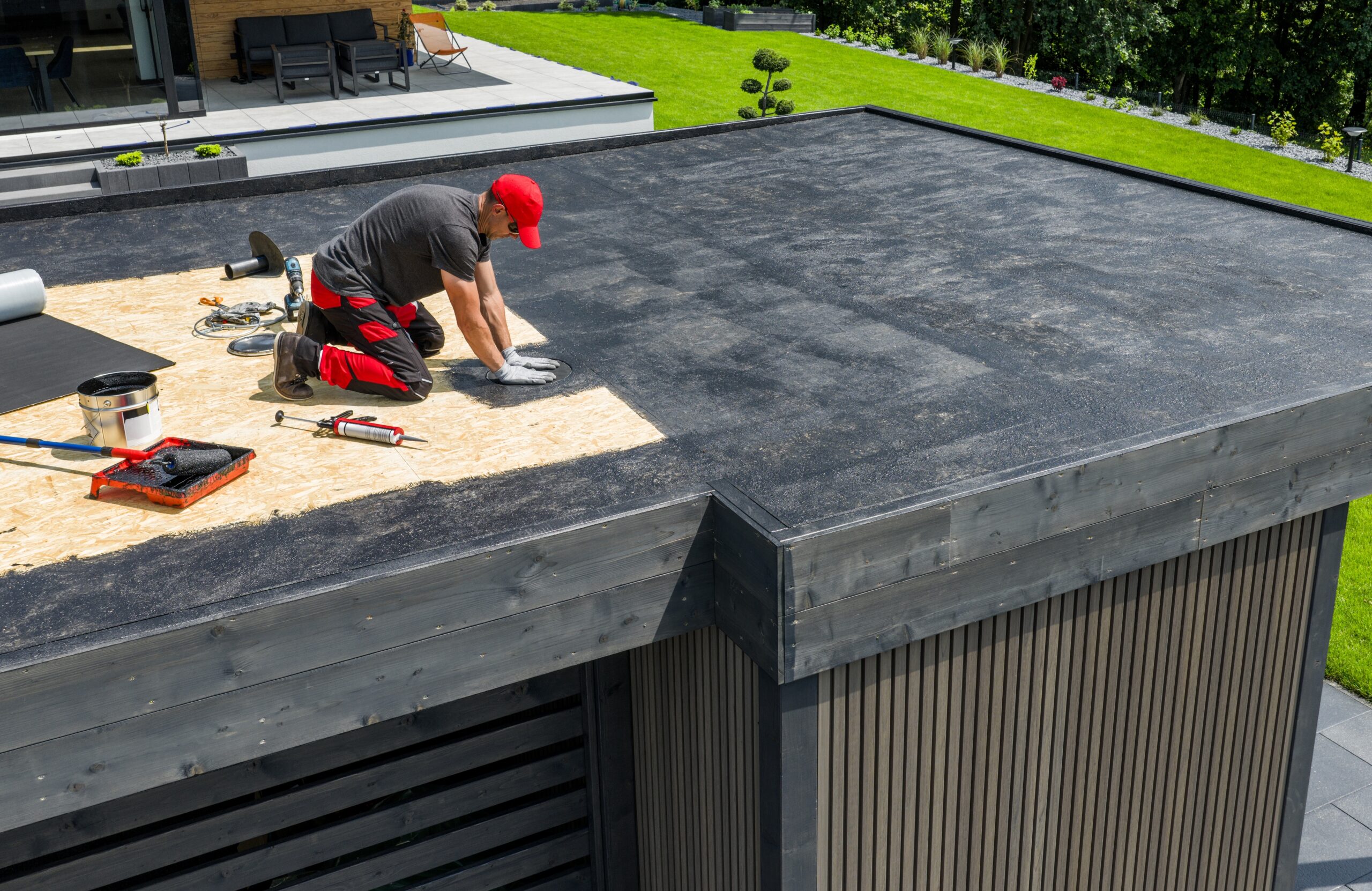 A roofer in a red cap installing a new black membrane on a residential flat roof, illustrating the comparison between flat roof repair vs replacement cost.