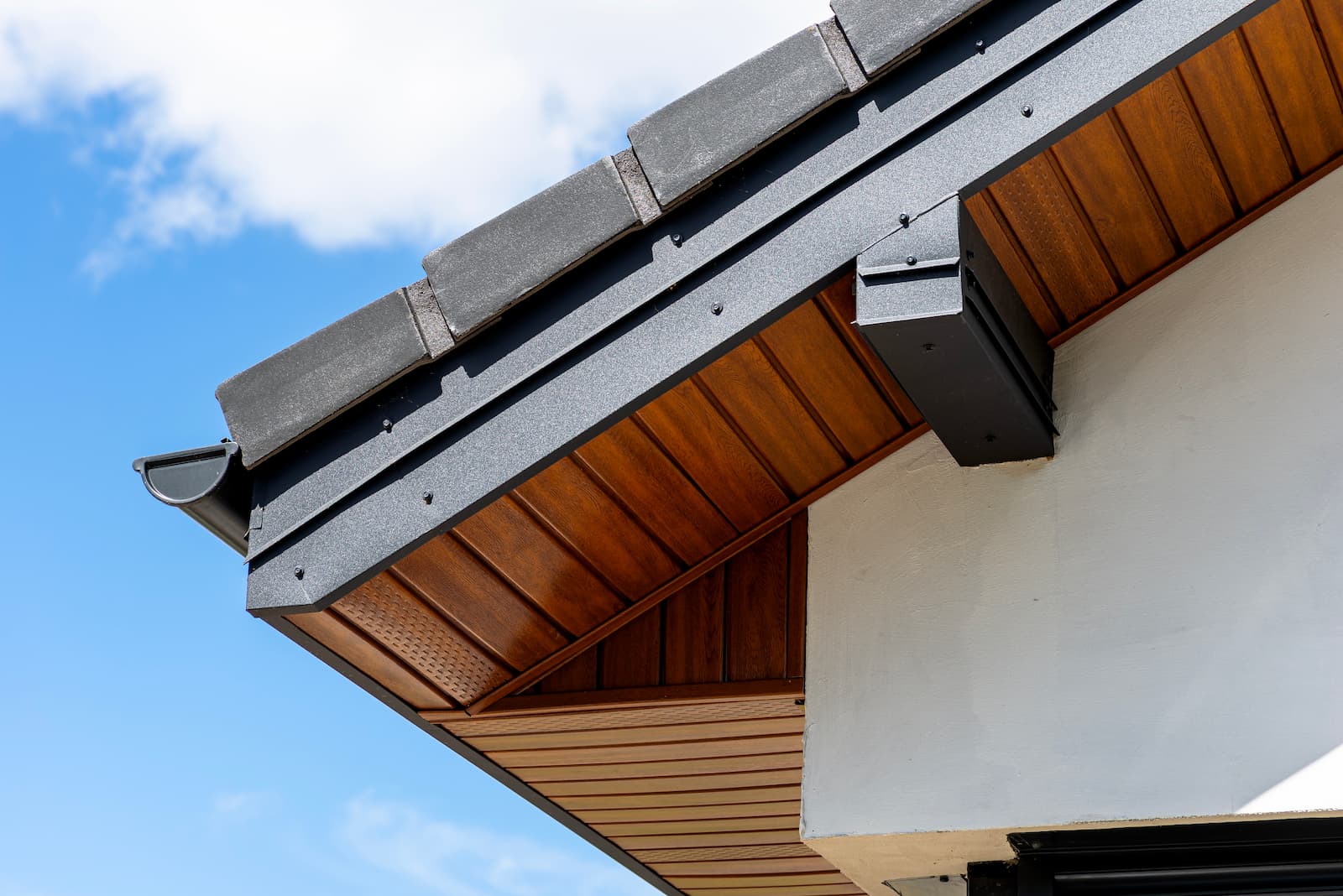 A low-angle shot of a modern roof corner featuring clean dark grey shingles, dark grey fascia board with a gutter, and polished wood-look soffit panels against a blue sky.