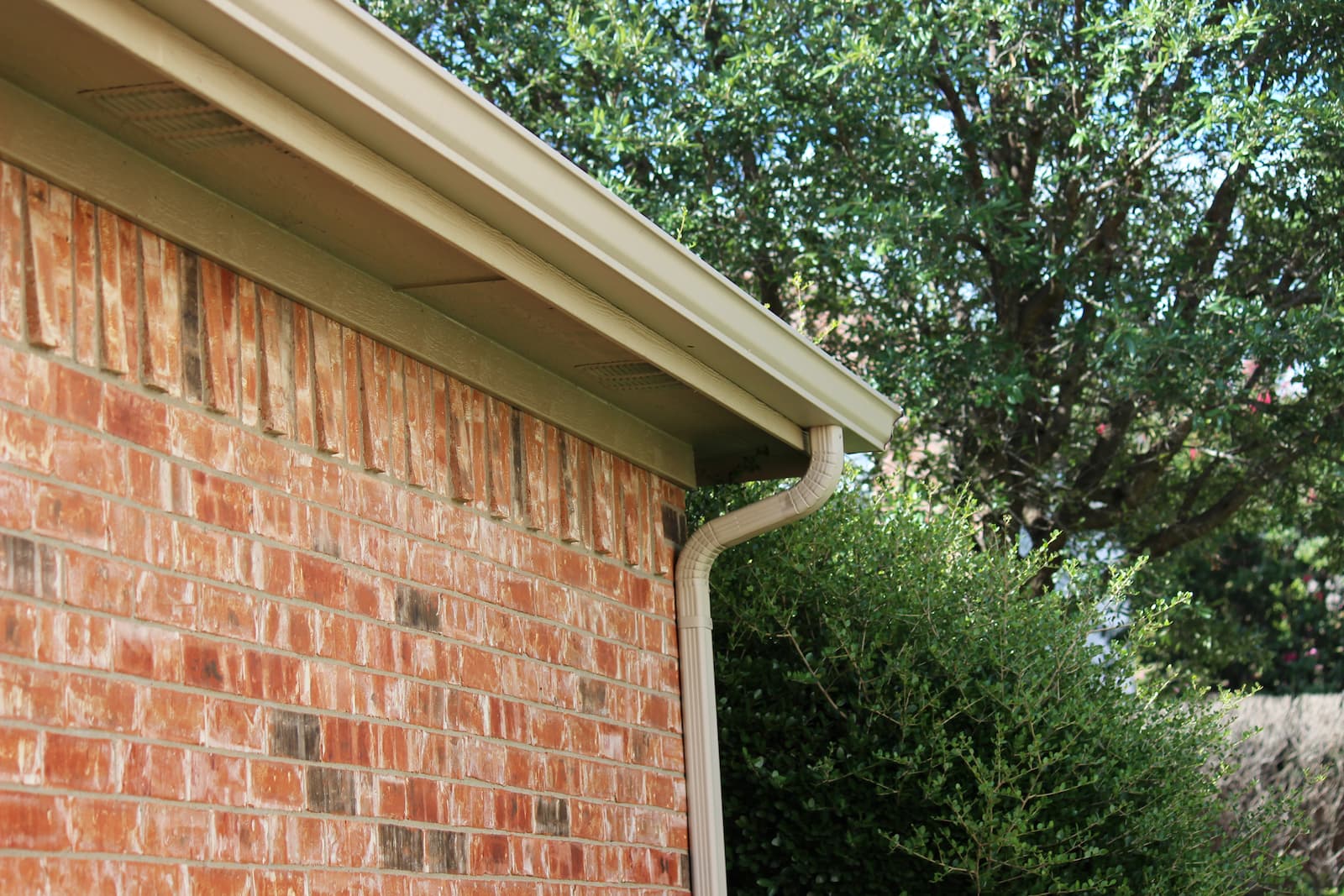 A close-up of a tan seamless gutter and downspout installed along the roofline of a red brick house with green trees in the background.