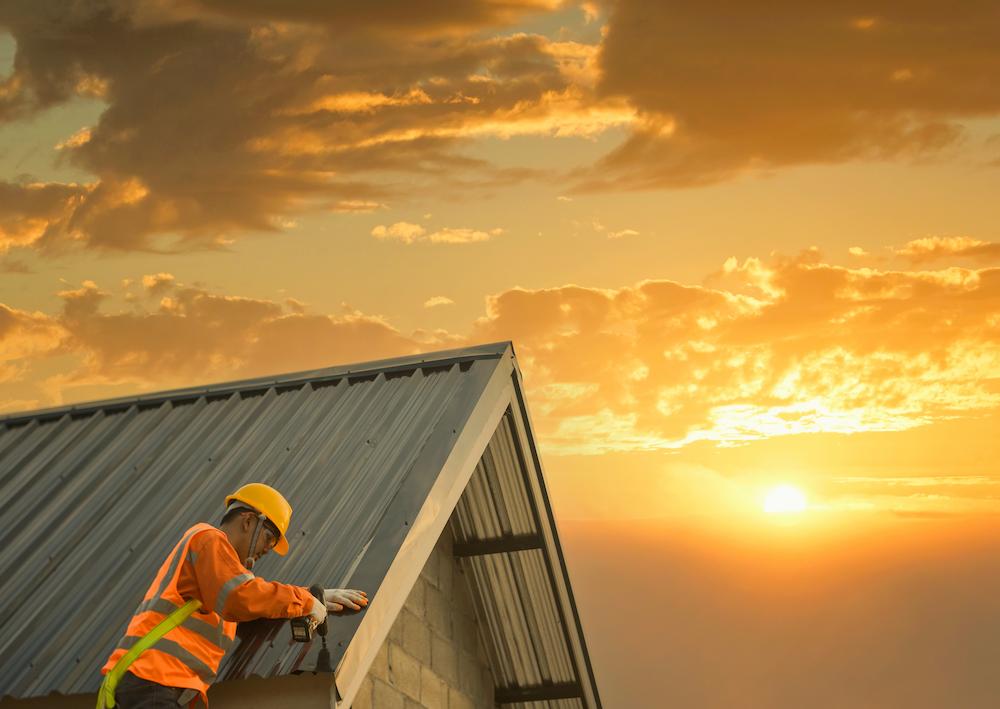Construction worker in a yellow hard hat and safety vest installing a metal roof at sunset.